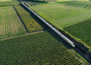 Aerial view of a sleek passenger train traveling through lush green fields with well-organized rows of crops, showcasing efficient transportation and agriculture.