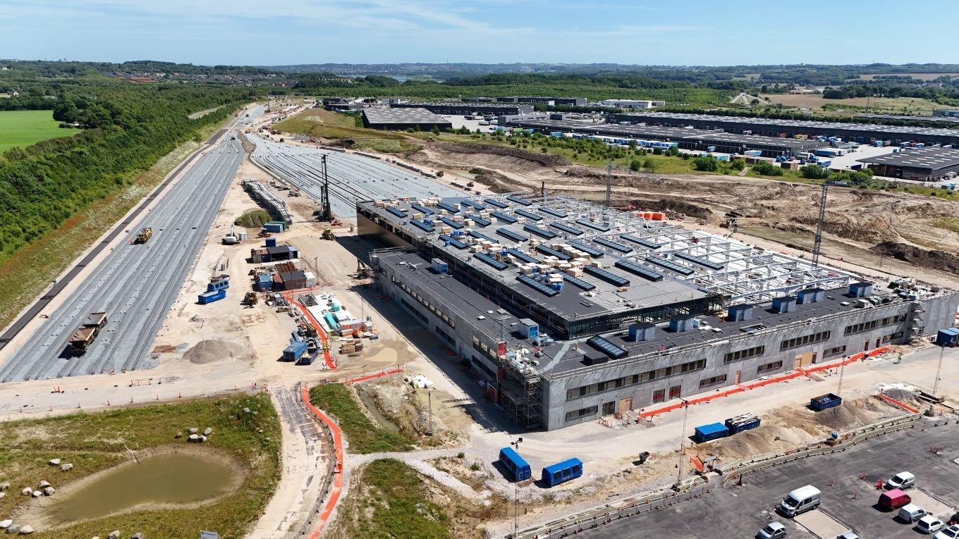 Aerial view of a DSB’s new rail repair and maintenance facility in Aarhus. Adjacent to the building, there are long stretches of concrete paths, construction equipment, and vehicles. Surrounding the site are green fields, small bodies of water, and other industrial buildings in the background. The sky is clear with light clouds visible.