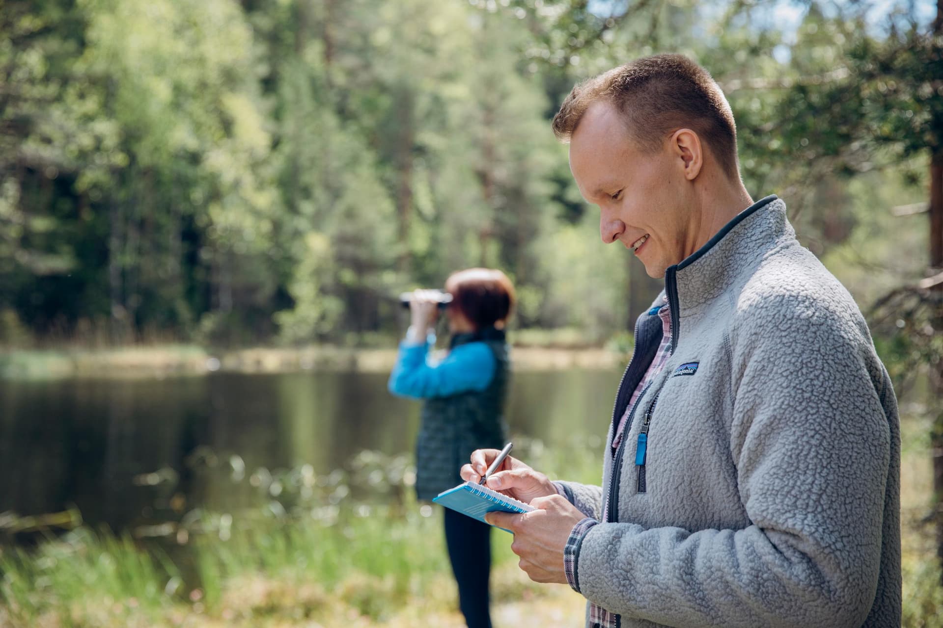 RFI brand photoshoot in Espoo, Finland. Environmental Specialist taking notes while a colleague is standing by the water and looking at birds with binoculars in Nuuksio National Park, Finland.
